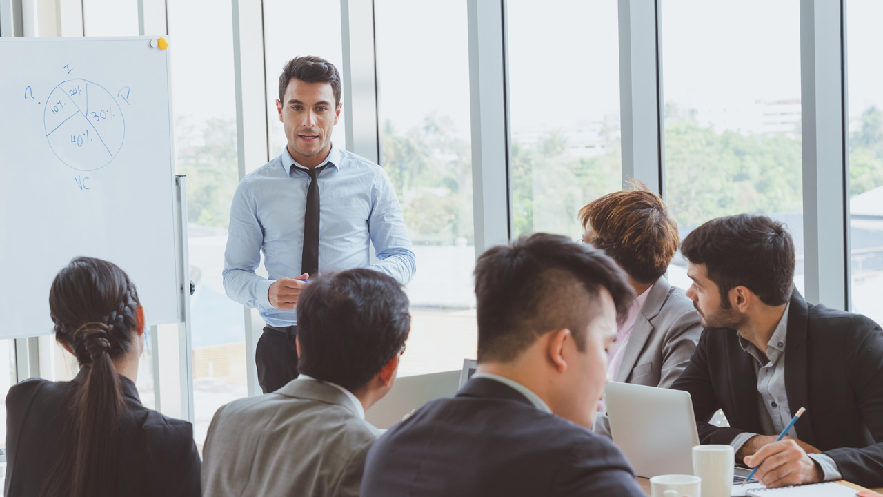 A man in a blue shirt and tie speaking to a group of people seated around a table in a meeting room